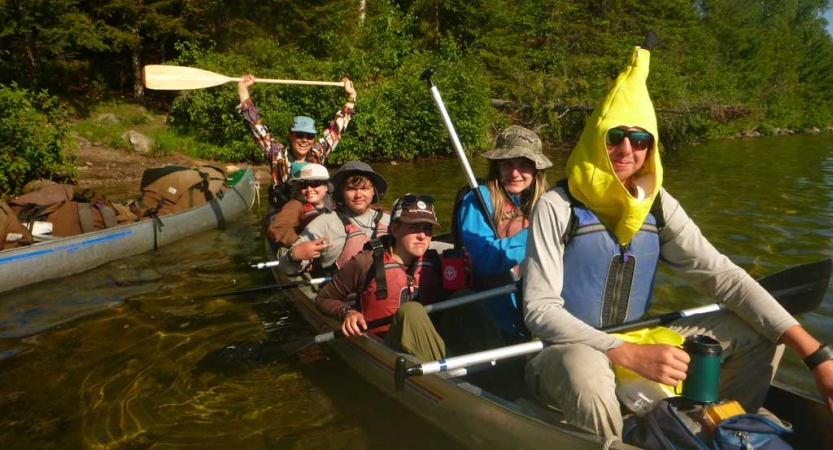 A group of students wearing lifejackets sit in a canoe and smile for the photo. One of them is wearing a banana costume. 